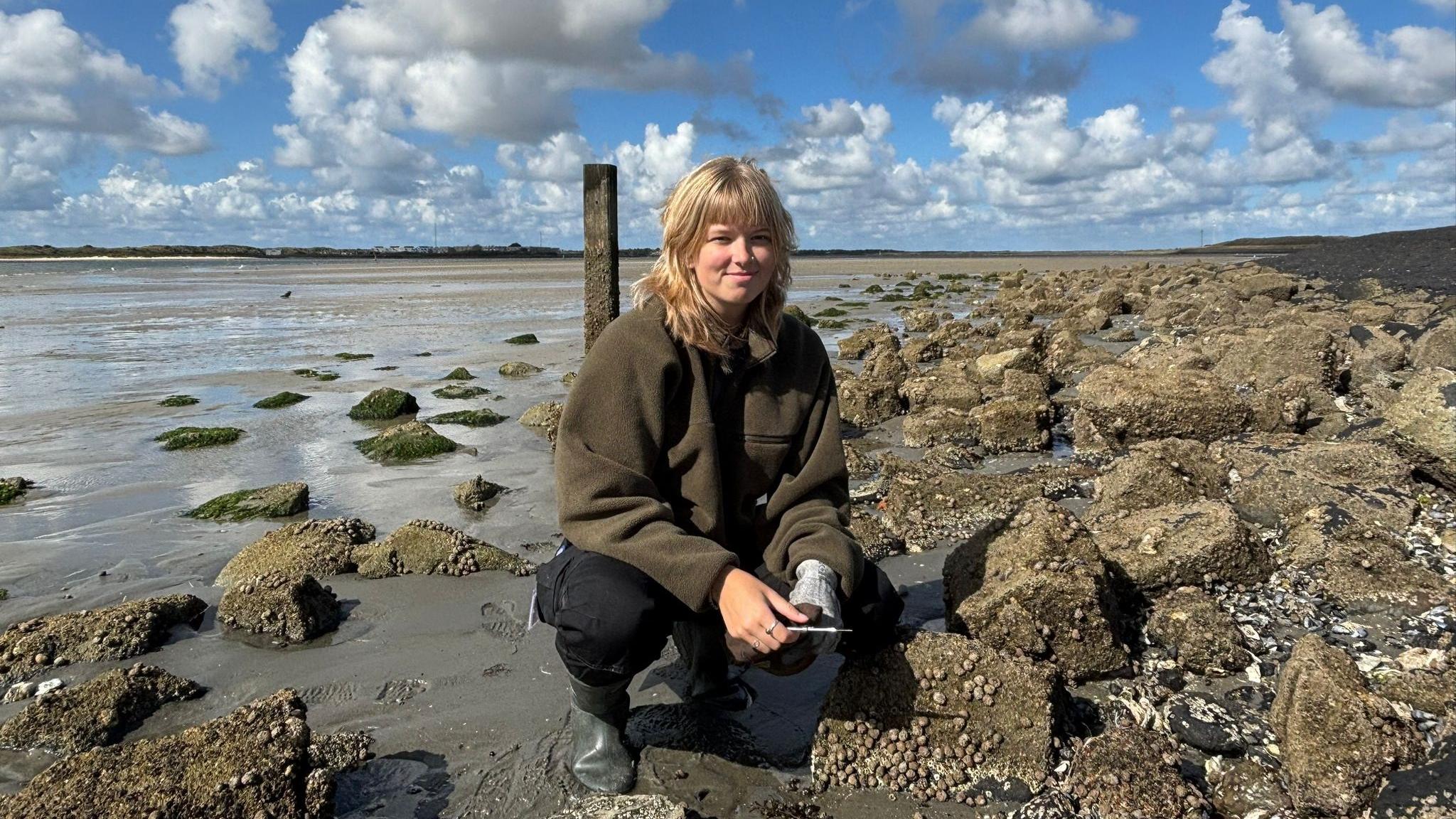 Rosalinde van Ooijen crouching on a rocky shore during low tide, surrounded by barnacle-covered rocks and patches of green seaweed. She is wearing a green jacket, black pants, and rubber boots, with a partly cloudy sky and tidal flats stretching into the distance.