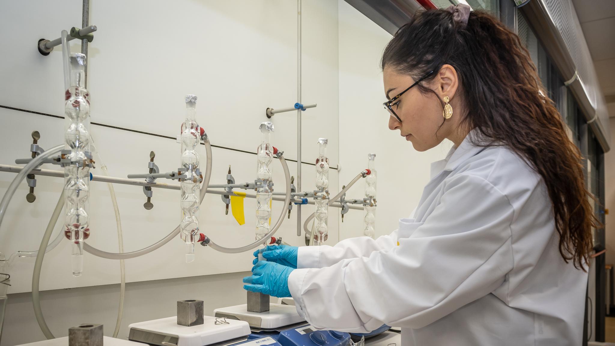 A woman working in a laboratory. In front of her a bench with four scale-like machines, with a hollowed out iron cube on each where a sample can be put into. Above each cube a glass tubelike structure, where samples will come from.