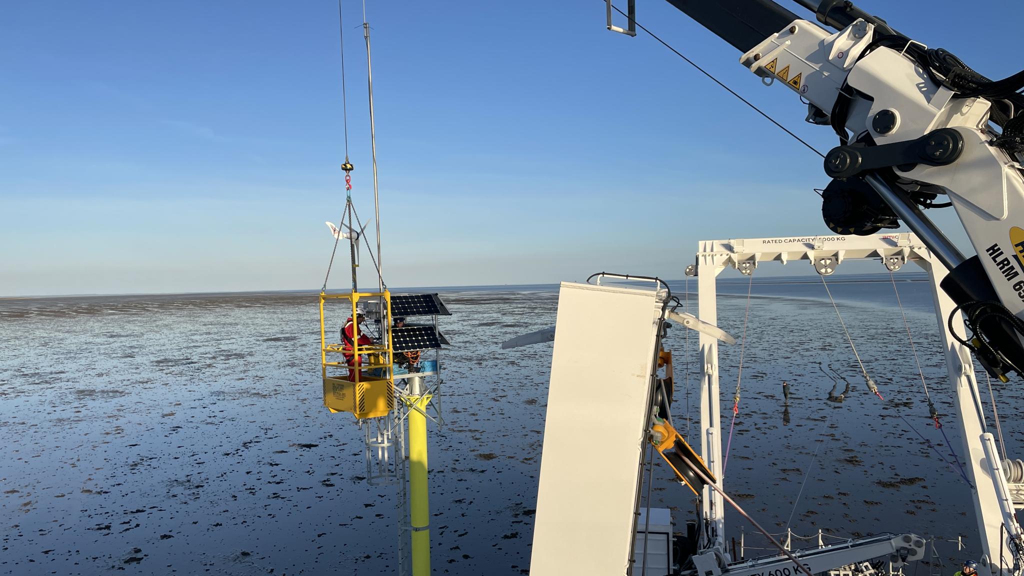 Een kraan op het dek van een schip houdt een hijskraan met een persoon erin vast naast een ijzeren paal die in de zeebodem is geplaatst. De persoon installeert een zonnepaneel bovenop de paal.