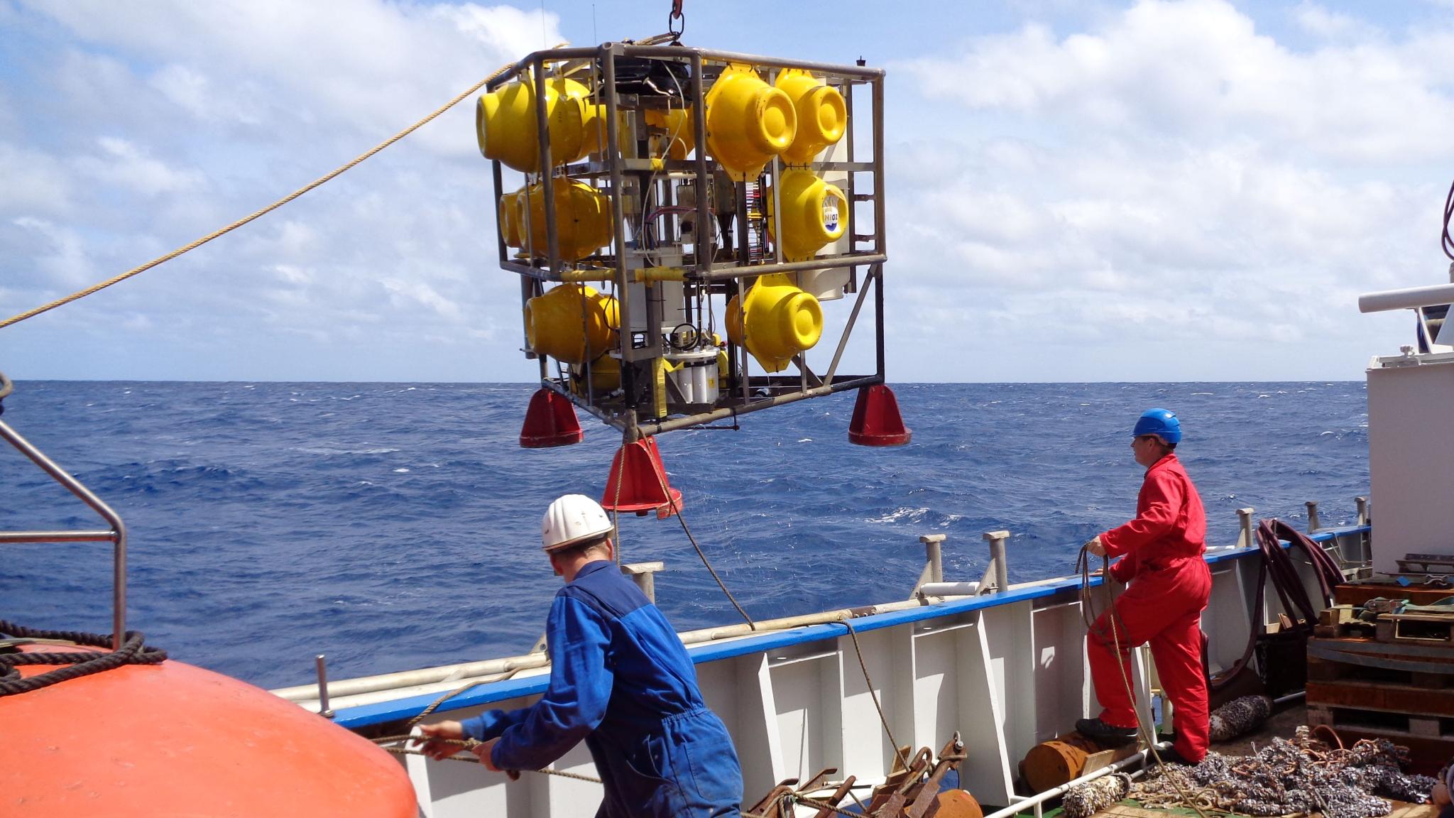Two people on the deck of a research vessel. An iron frame with large yellow balls in it is lifted from the deck to be lowered into the ocean
