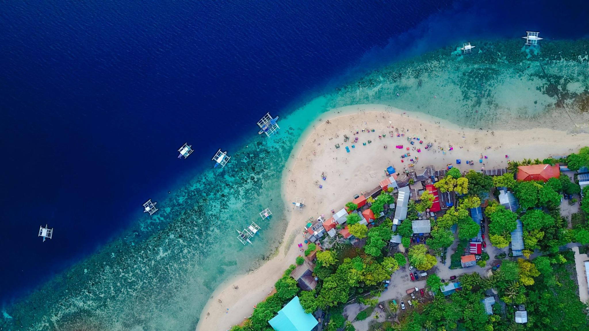 Aerial photo of a tropical beach with turquoise water, small boats along the coast and colourful parasols, surrounded by lush green trees and small houses (photo: Tirachard Kumtanom)