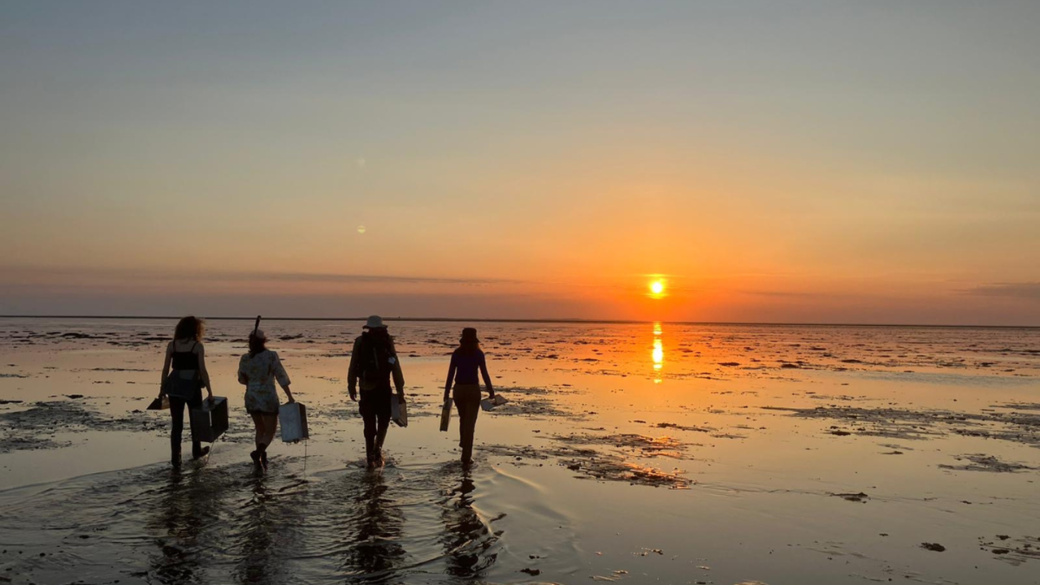 Four people walk across a shallow beach at sunset, their silhouettes reflected on the wet sand.