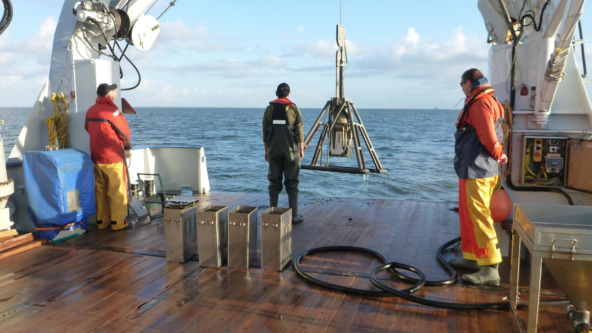 Three people on the back of the deck looking out over the Wadden Sea. One person is standing in the middle behind a box core. Photo by Bianka Rasch.
