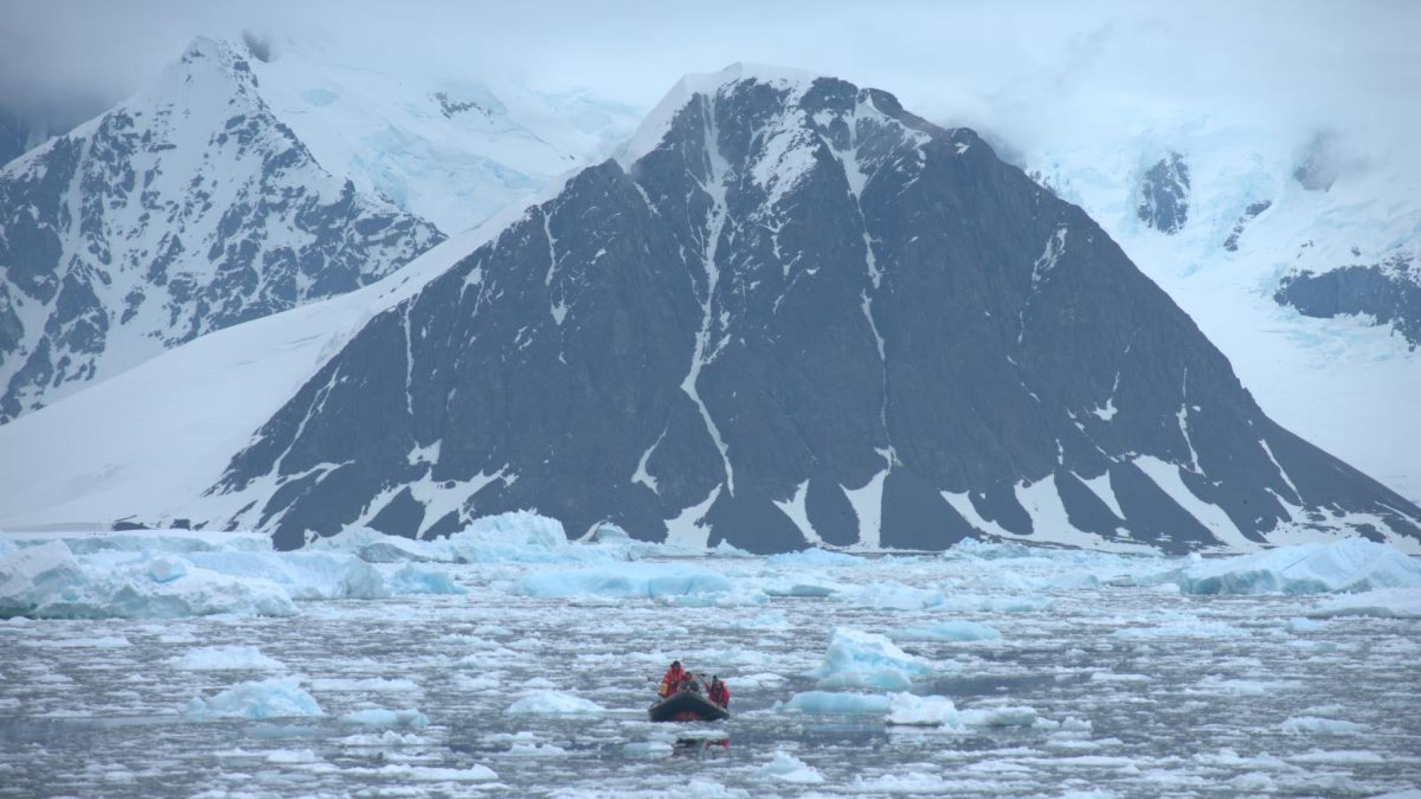 A small inflatable boat carrying researchers moves through icy water scattered with floes, heading toward a towering, snow-covered mountain with dark rock faces under a cloudy Antarctic sky.