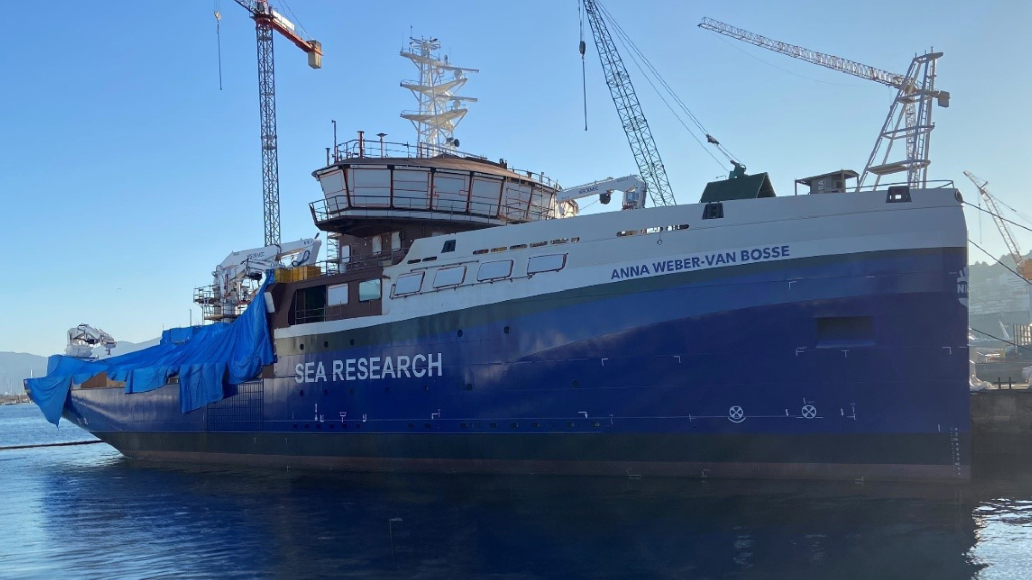 The ship RV Anna Weber van Bosse being under construction, seen from the side. The back deck is covered with palstic, and there are 3 cranes behind the ship lifting supplies on deck.