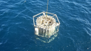 A CTD rosette sampler being recovered from the sea, partially lifted out of the water on a cable. The frame holds multiple vertical sampling bottles arranged in a circular structure.