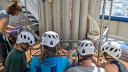 Group of researchers wearing helmets and gloves collecting water samples from the Niskin bottles on a CTD rosette sampler aboard a research vessel. The cylindrical bottles are mounted vertically in a metal frame, with tubing used to draw water into sample containers.