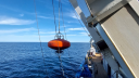 The side of a ship (the RV Pelagia) recovering a red/orange mooring device from the water.