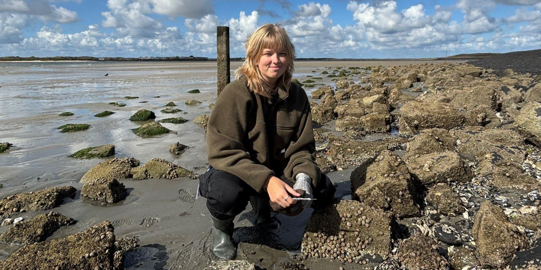 Rosalinde van Ooijen crouching on a rocky shore during low tide, surrounded by barnacle-covered rocks and patches of green seaweed. She is wearing a green jacket, black pants, and rubber boots, with a partly cloudy sky and tidal flats stretching into the distance.