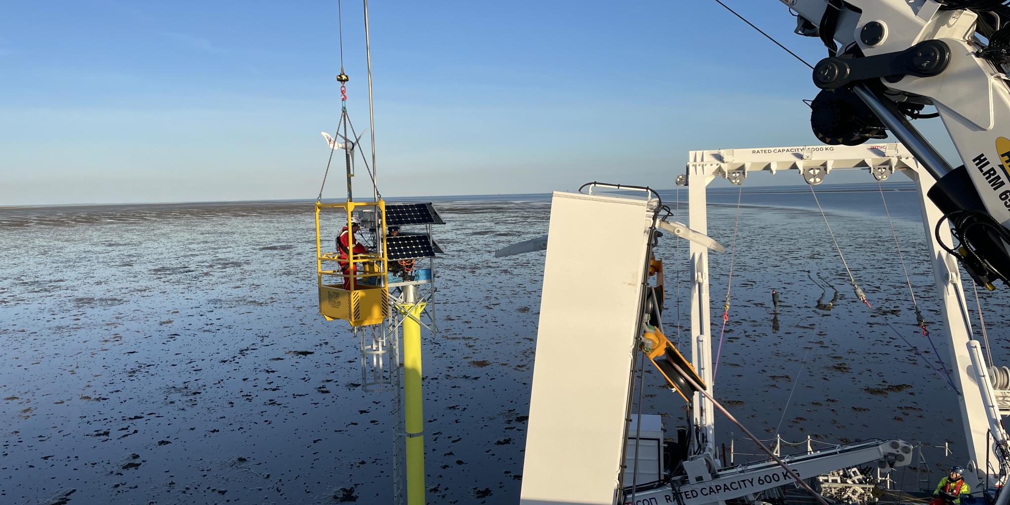 Een kraan op het dek van een schip houdt een hijskraan met een persoon erin vast naast een ijzeren paal die in de zeebodem is geplaatst. De persoon installeert een zonnepaneel bovenop de paal.