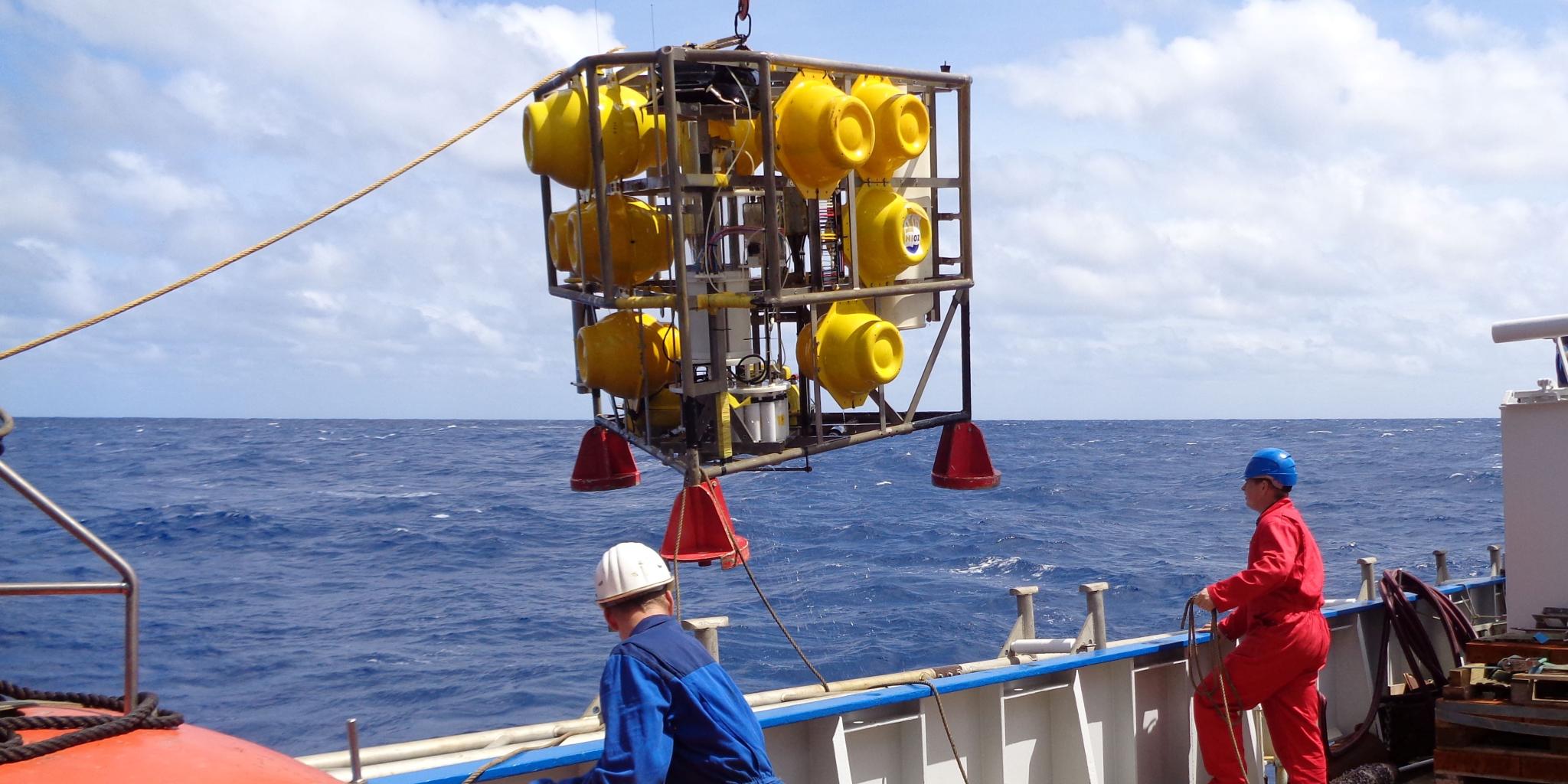 Twee mensen op het dek van een onderzoeksschip. Een ijzeren frame met grote gele ballen erin wordt van het dek getild om in de oceaan te worden neergelaten.