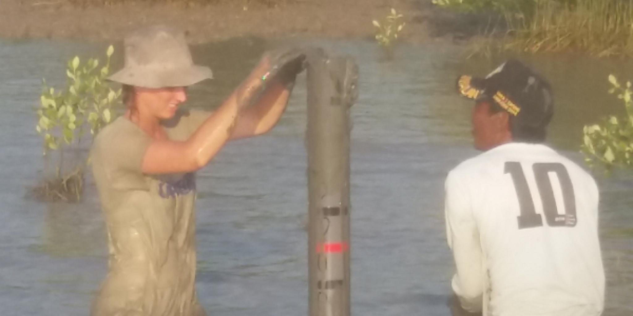 Two people standing knee-deep in muddy water installing a vertical measuring tube or pole; one is wearing muddy work clothes and a hat, and the other is wearing a white shirt with the number 10 on the back.