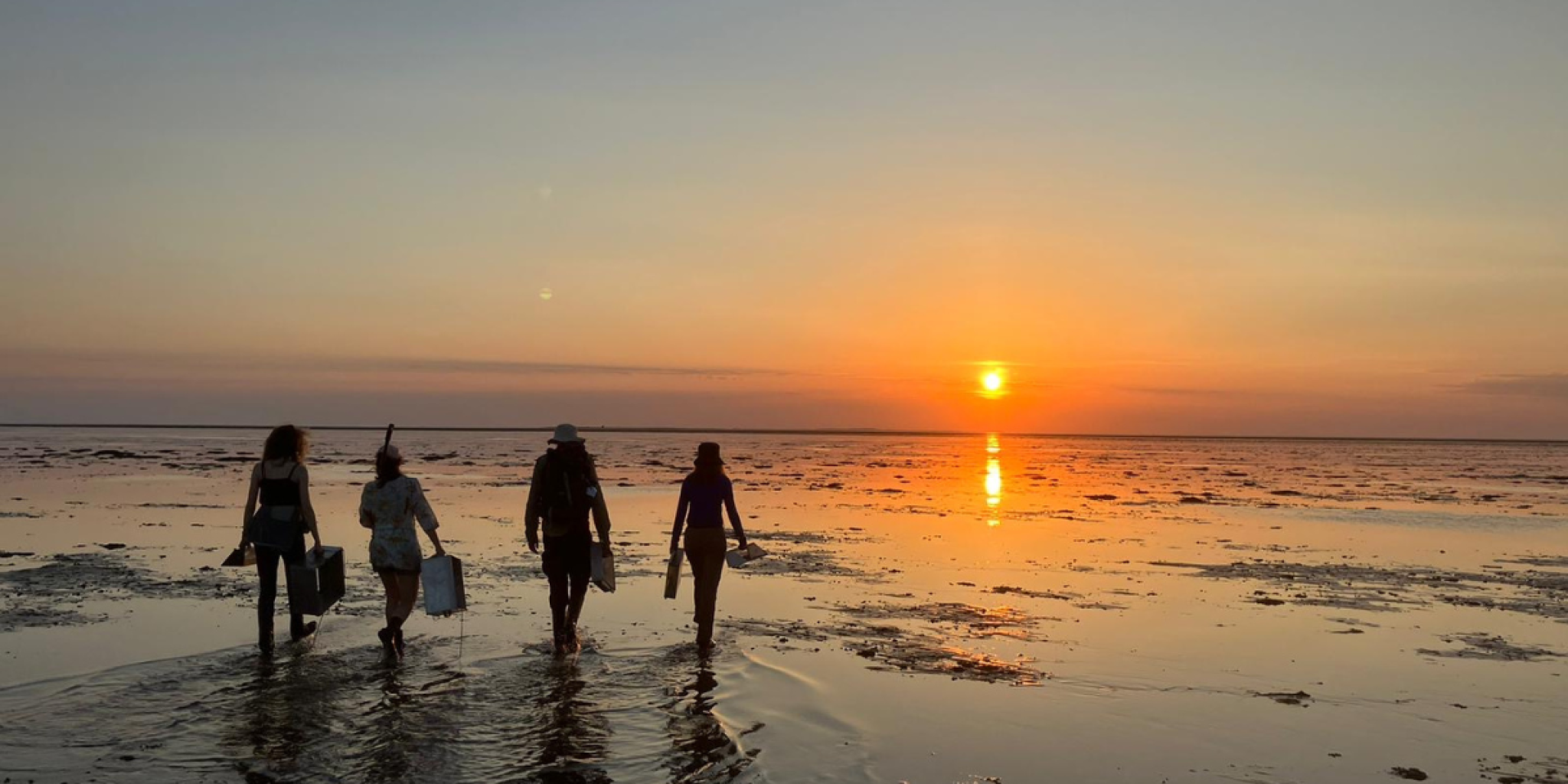 Four people walk across a shallow beach at sunset, their silhouettes reflected on the wet sand.
