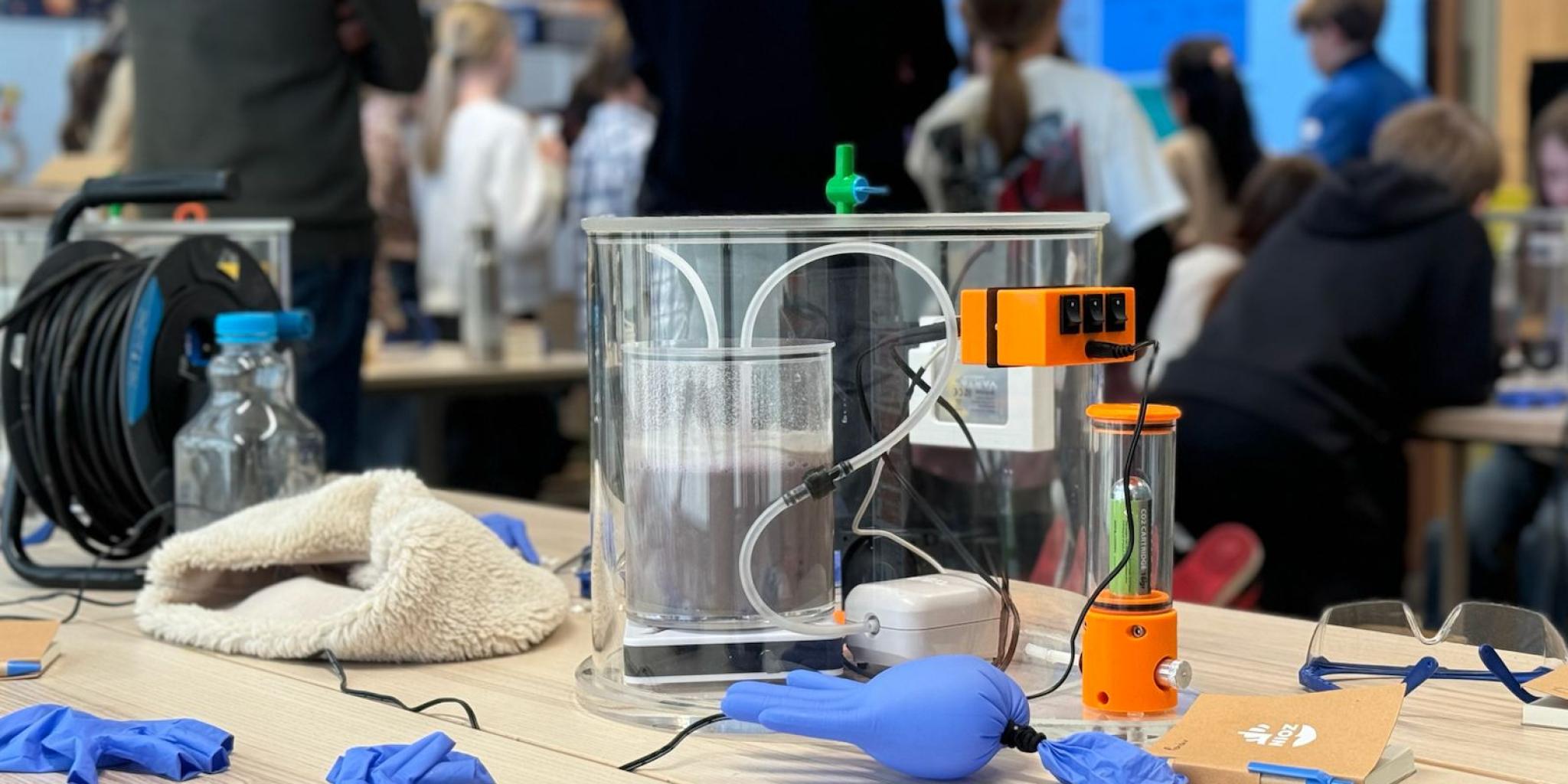 A demonstration setup with a clear tank and sensors on a table, surrounded by lab gloves and equipment, while people watch in the background.
