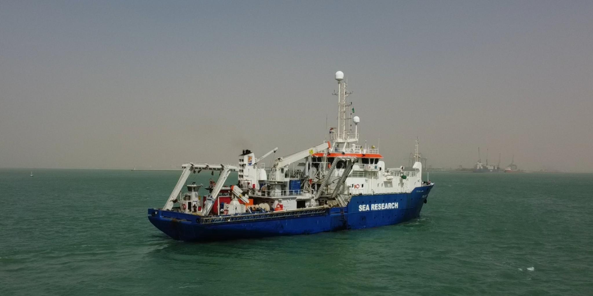 Een blauw-wit onderzoeksschip met het opschrift "SEA RESEARCH" varend op kalm groen water.