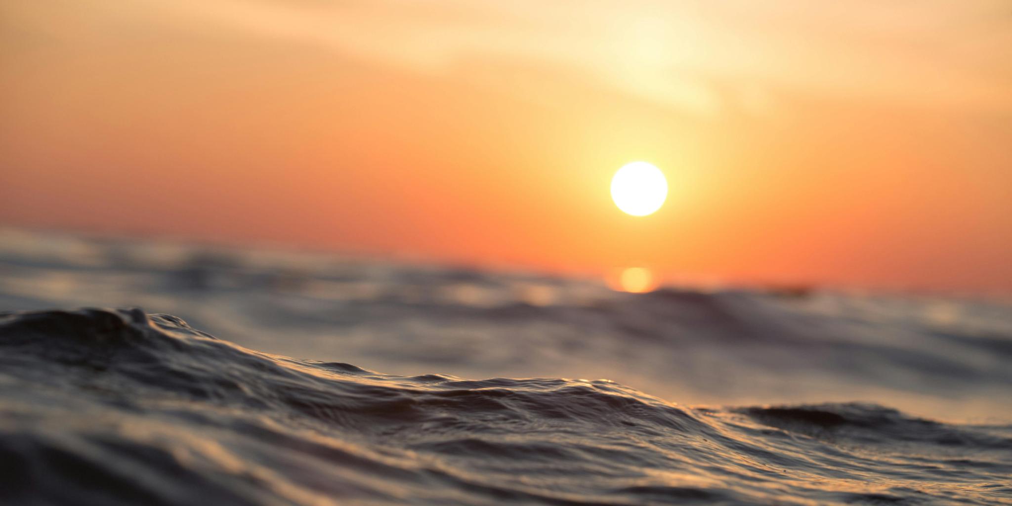 Zonsondergang op zee in tinten oranje en geel. Op de voorgrond zie je een watergolf.
