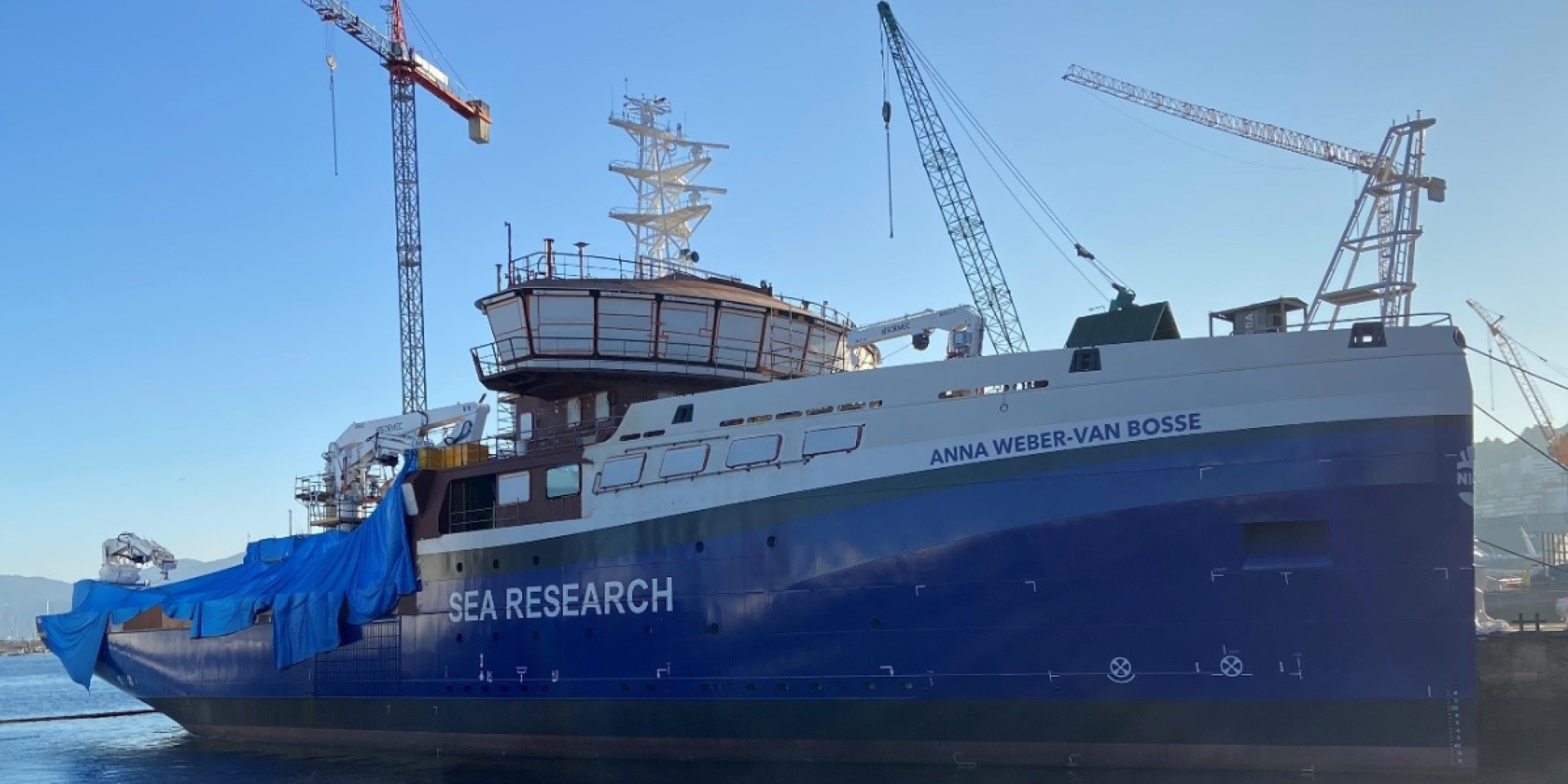 Het schip RV Anna Weber van Bosse in aanbouw, gezien van de zijkant. Het achterdek is bedekt met plastic, en er zijn 3 kranen achter het schip die benodigdheden op het dek hijsen.