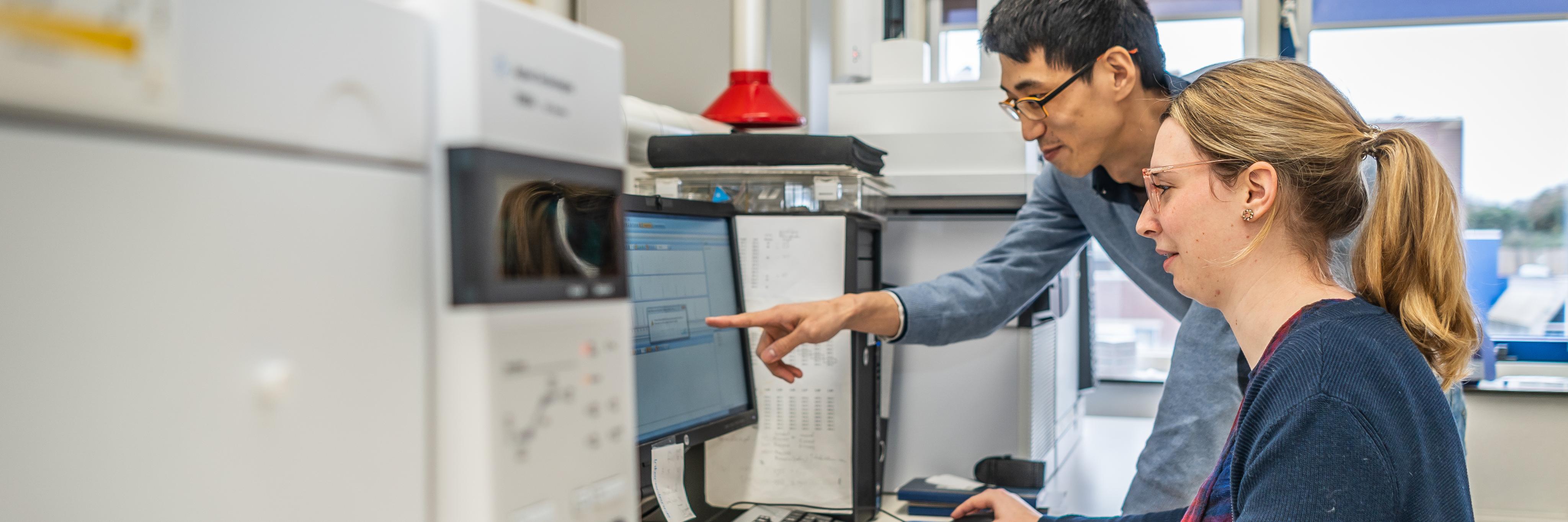 Two researchers looking at a computer, one pointing at the screen. The computer is connected to several laboratory machines, looking like big grey boxes atop a working bench.