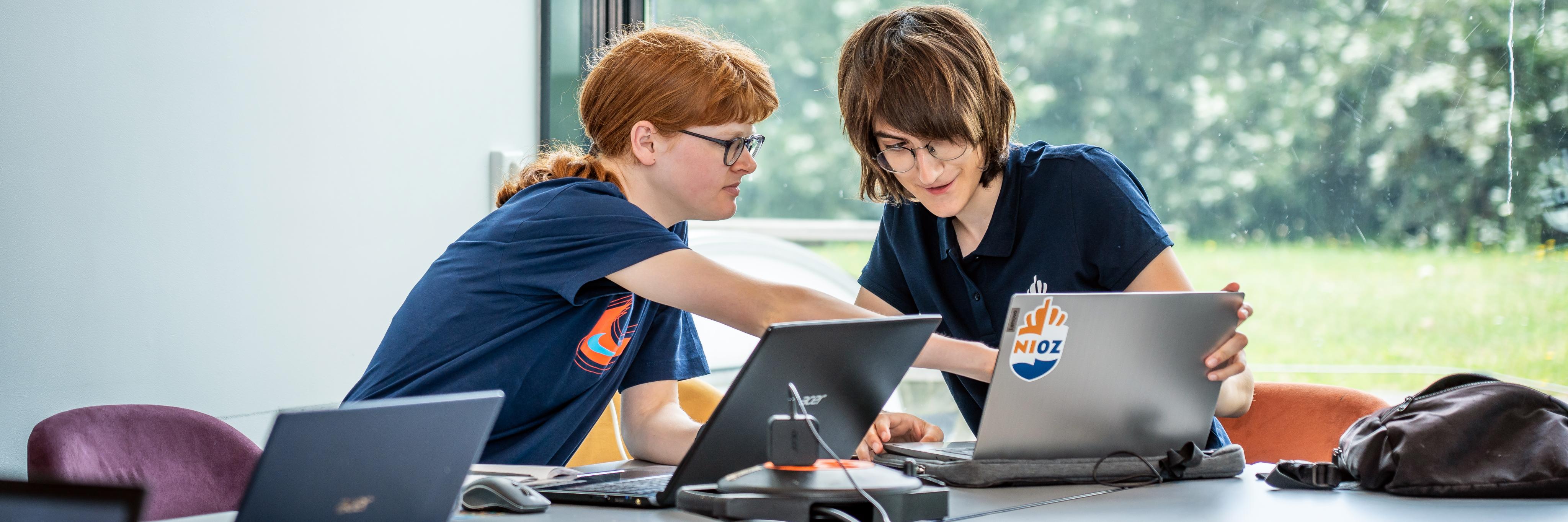 Two students sitting at a table with laptops. They are researching something and pointing at the screen of one of the laptops.
