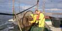 Two people in bright yellow waterproof gear and life jackets working at a fyke net setup in shallow coastal waters. They are on a small boat alongside large circular nets held up by wooden poles, used for fish sampling at the NIOZ research station Texel.