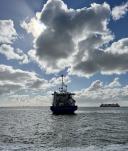 The RV Pelagia sailing off. The view is from the back. You can see a cloudy sky with sunshine.